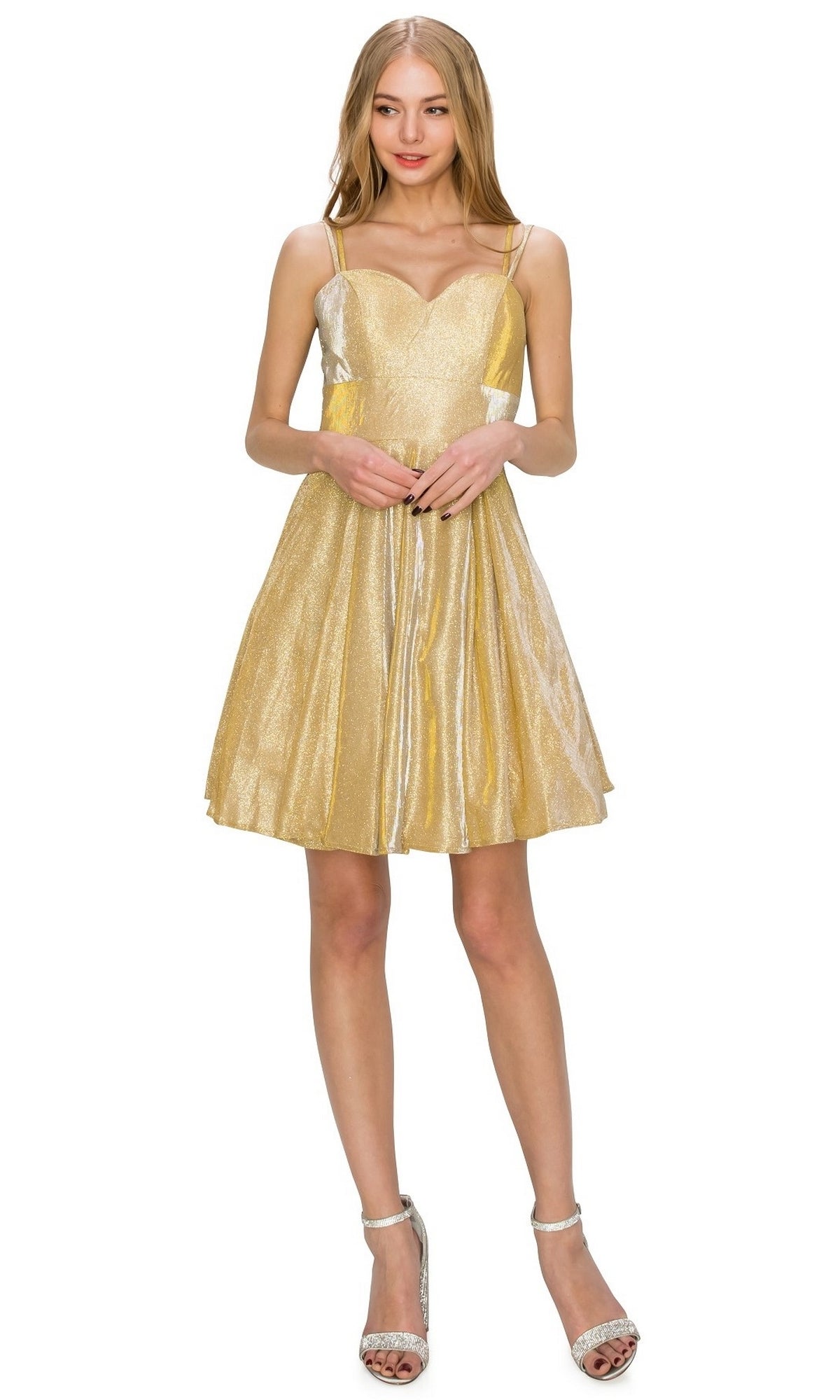 A woman wearing the Lace-Up Short Glitter A-Line Homecoming Dress 8011J—sleeveless, knee-length, metallic gold with thin straps—and silver high-heeled sandals stands against a white background.